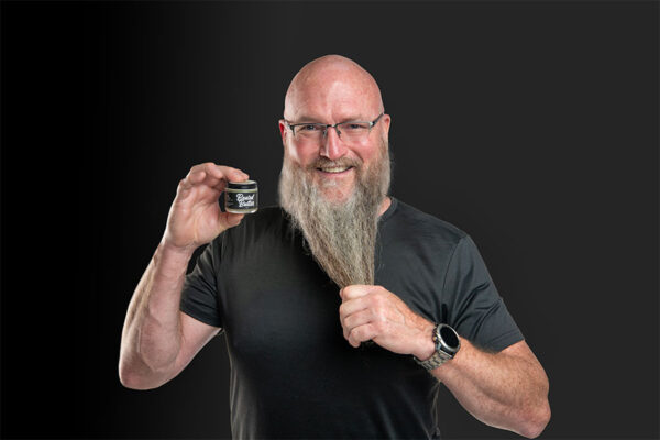 Man in black t-shirt and glasses smiling, pulling on his long beard while holding up a jar of Beard Butter beard care product.