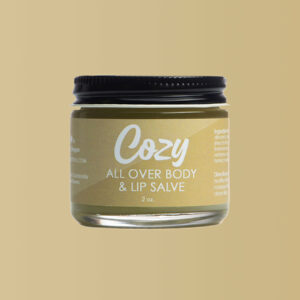 Close up image of Cozy All Over Body and Lip Salve against a neutral sand colored background.