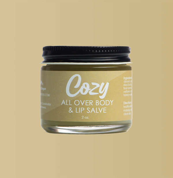 Close up image of Cozy All Over Body and Lip Salve against a neutral sand colored background.
