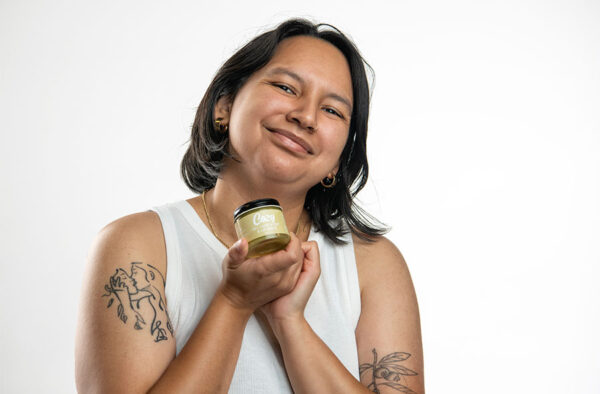 Headshot of woman with dark hair, smiling wearing a white tank top holding a jar of skin care salve against white background.