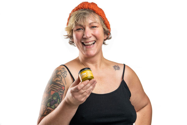 Smiling woman wearing orange knit cap and black shirt holding a jar of Glow all over body and lip salve.