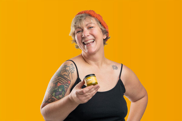 Woman smiling brightly against a mustard yellow background holding a jar of Glow all over body and lip care salve.