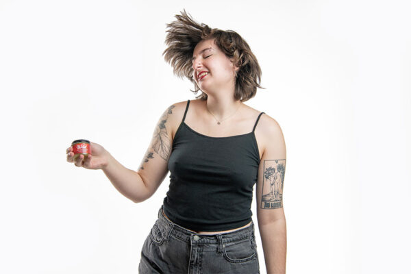 Young women with hair flying upwards, in dancing movement holding jar of Wild all over body and lip salve against white background.